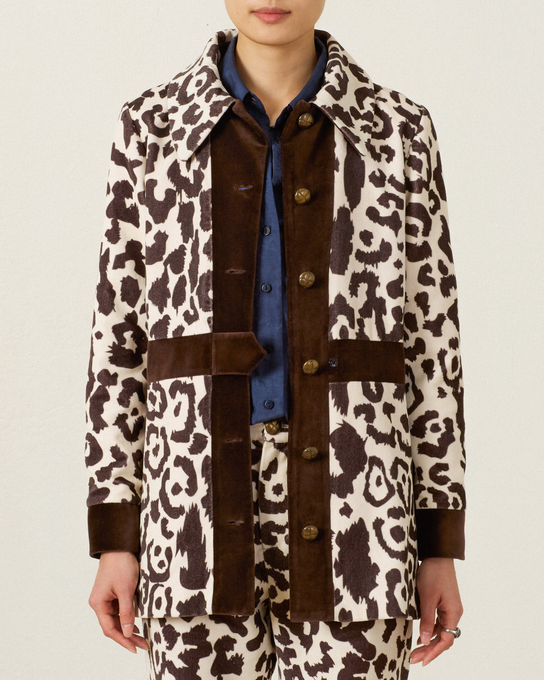Wearing the Lydia Leopard Steppe Jacket, featuring a vintage silhouette with brown cotton-velvet trim and a large collar, layered over a blue shirt and matched with leopard print pants, the person stands against a plain background.