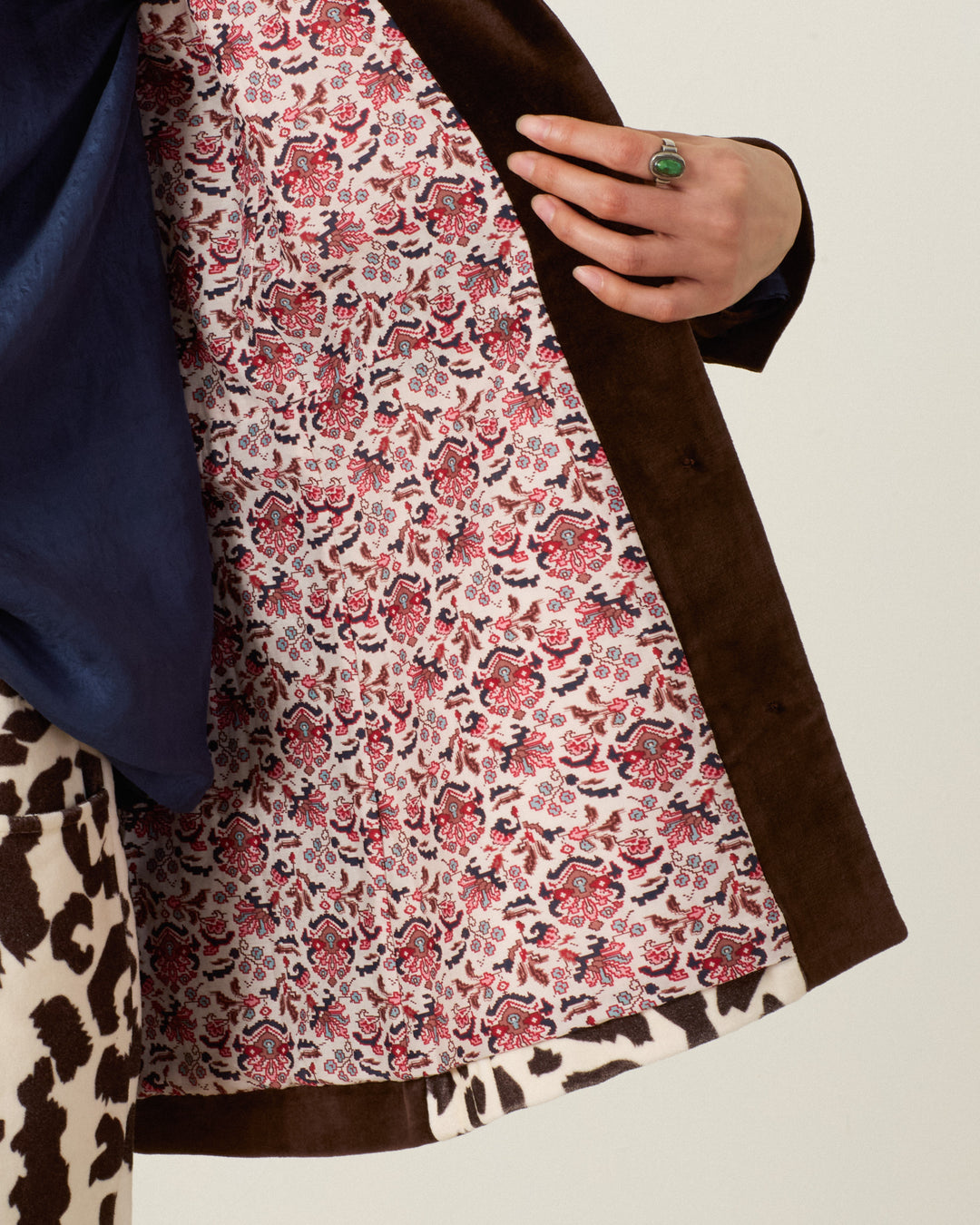 Wearing the Lydia Leopard Steppe Jacket, a person holds open the brown cotton-velvet coat to reveal its pink and white floral lining, paired with a blue top, leopard print pants, and a green stone ring.