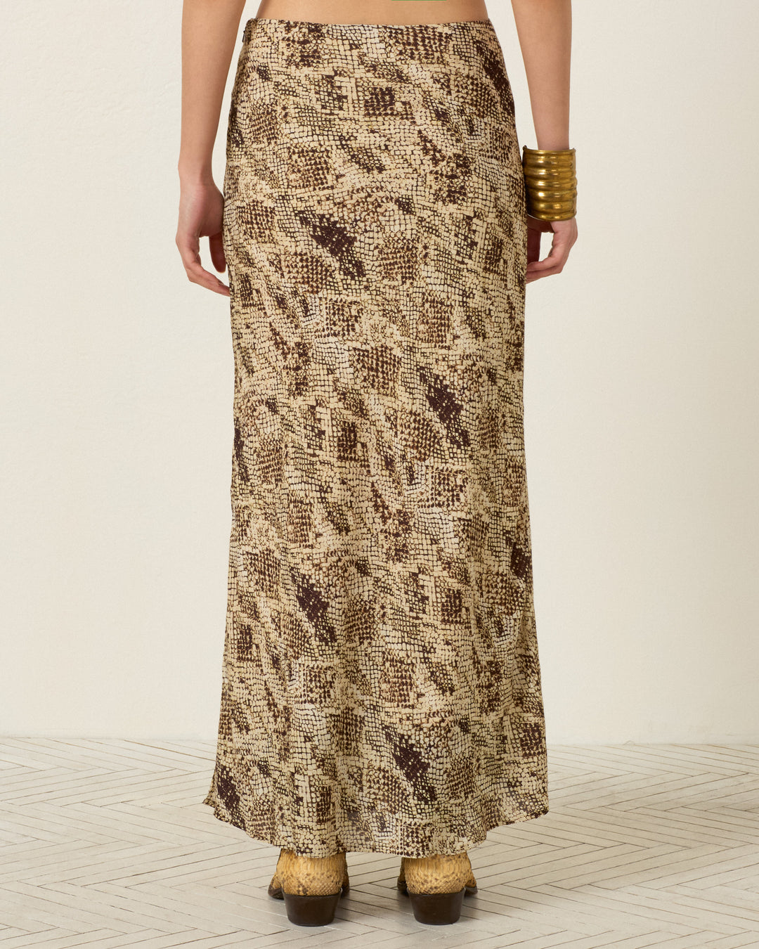 A person wearing the Lyra Lemon Snakeskin Skirt, styled with a gold bracelet and tan boots, is shown from the back against a light background.