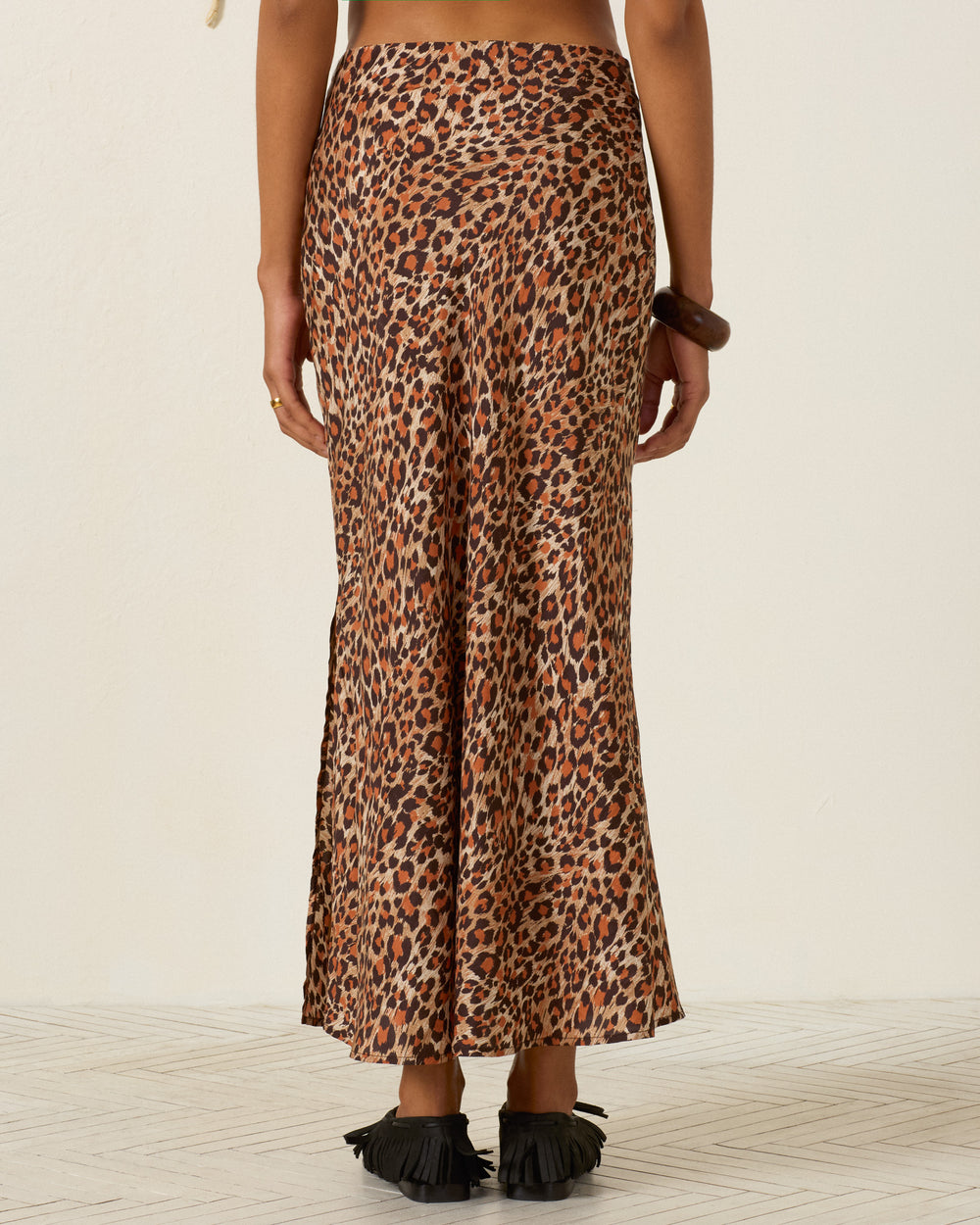 A person stands facing away, wearing the Lyra Leopard Trek Silk Skirt—a high-waisted, ankle-length bias cut silk skirt in a leopard print—paired with black fringed sandals against a plain light background.