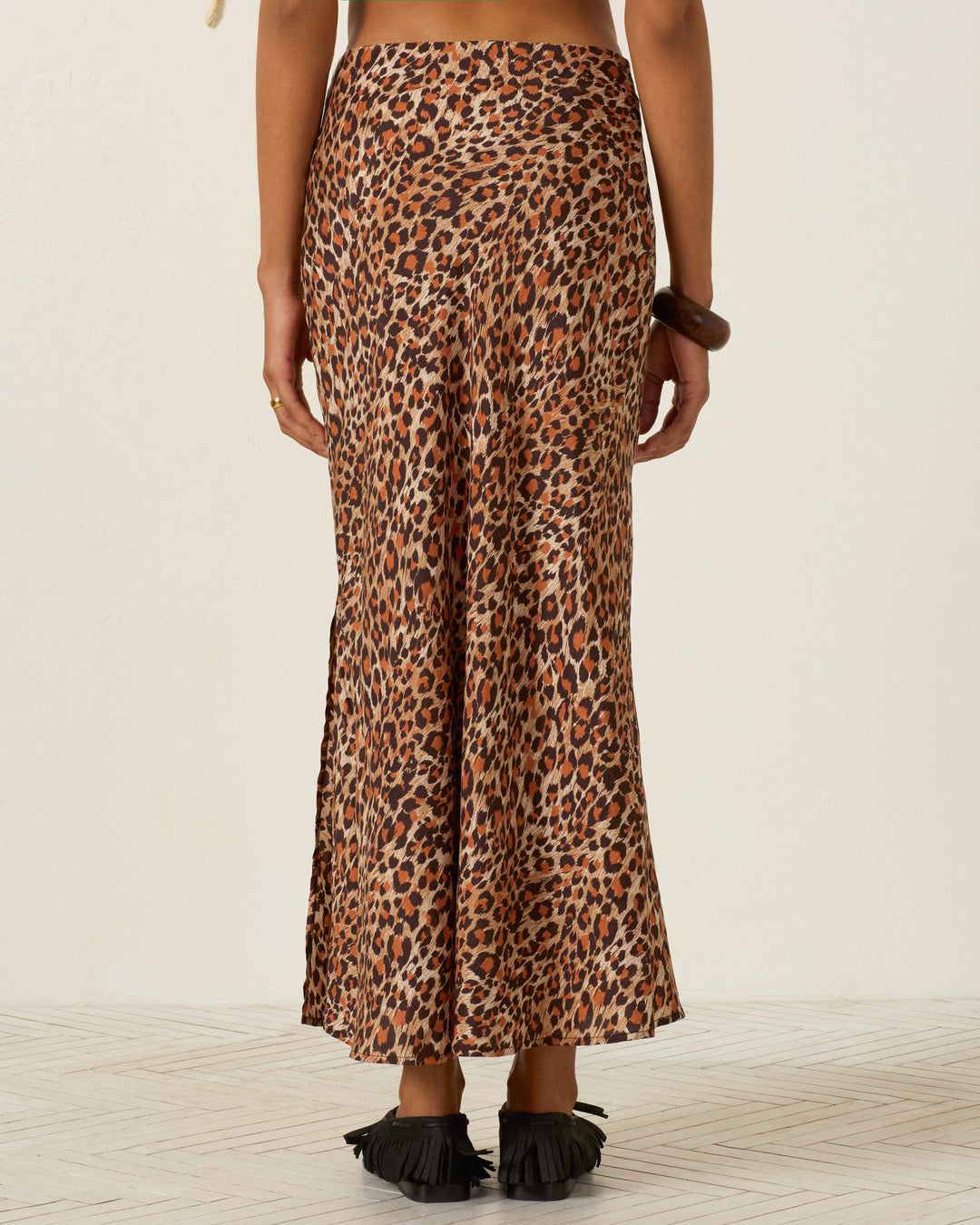 A person stands facing away, wearing the Lyra Leopard Trek Silk Skirt—a high-waisted, ankle-length bias cut silk skirt in a leopard print—paired with black fringed sandals against a plain light background.