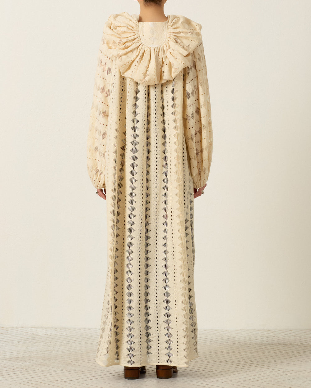 A person stands with their back to the camera, wearing the Marcie Savanna Lattice Dress—a long, patterned cream dress with a large ruffled collar and long sleeves adorned with delicate geometric lace detailing.