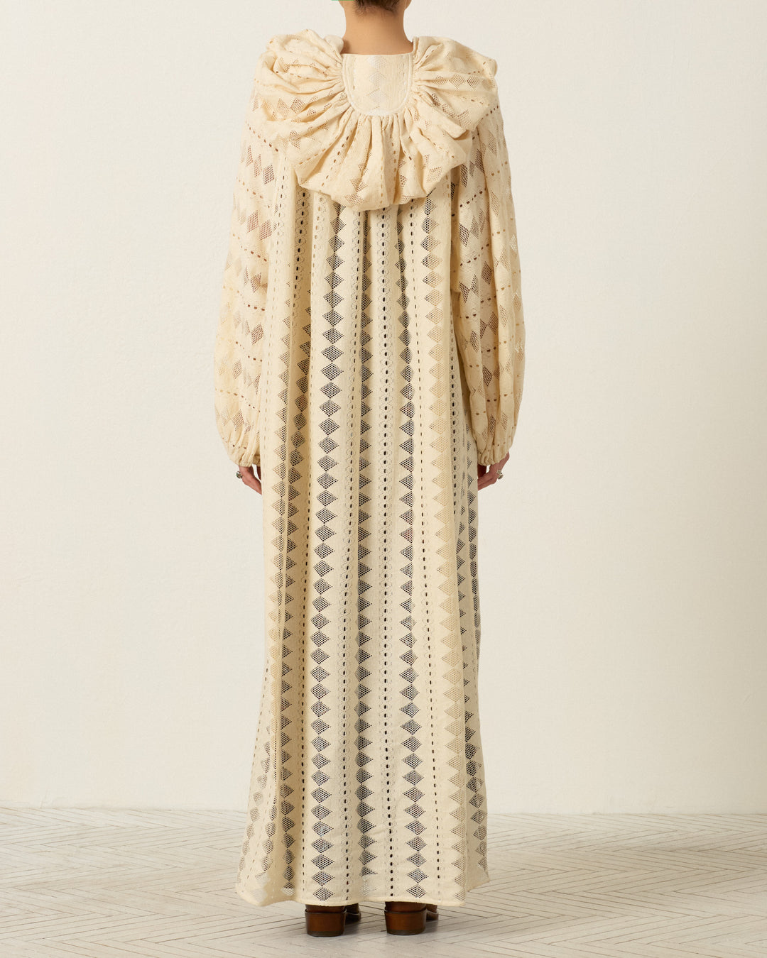 A person stands with their back to the camera, wearing the Marcie Savanna Lattice Dress—a long, patterned cream dress with a large ruffled collar and long sleeves adorned with delicate geometric lace detailing.