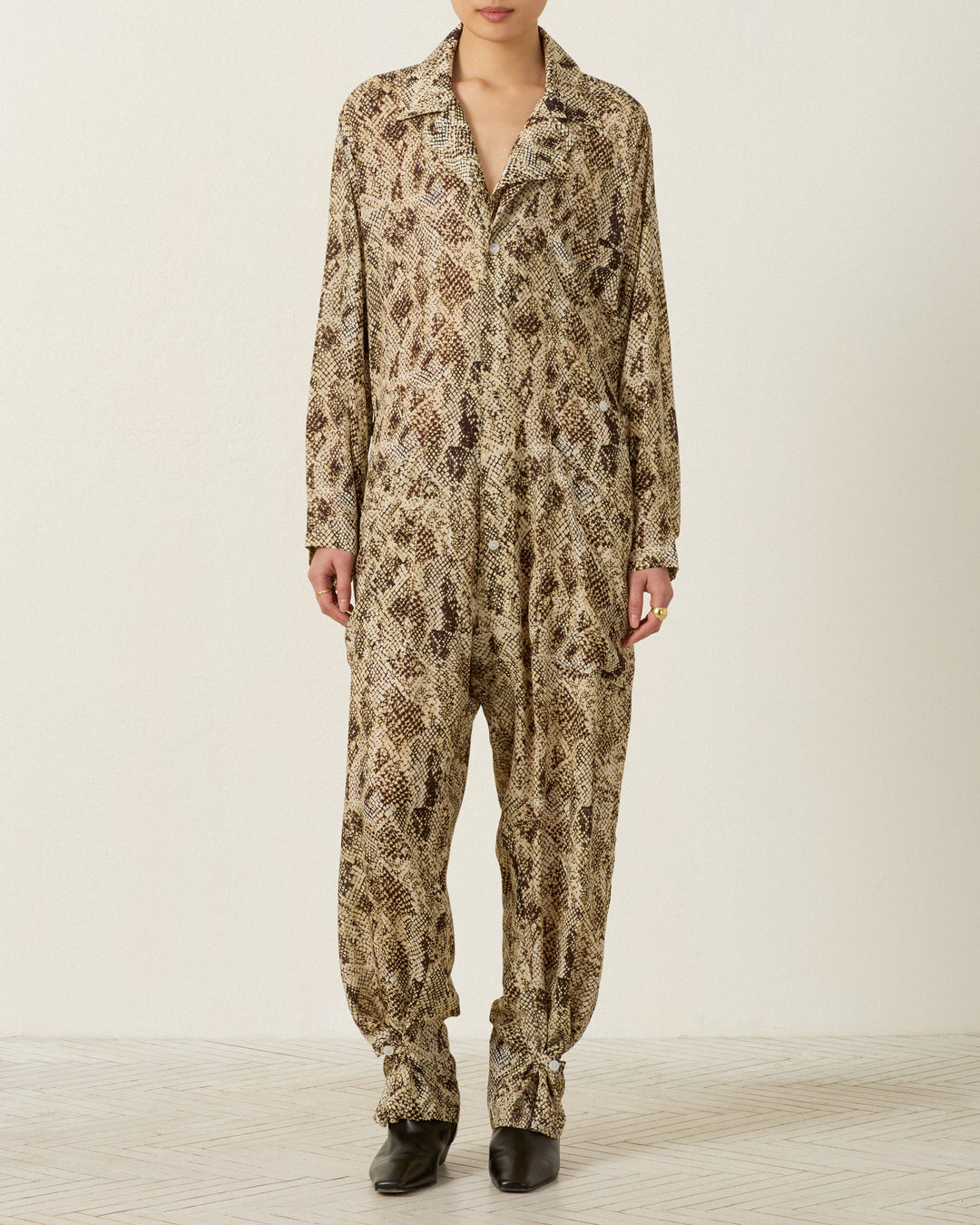 A person stands against a plain wall wearing the Miles Lemon Snakeskin Jumpsuit—a long-sleeved, button-up workwear-inspired piece featuring a brown and beige snakeskin pattern—paired with black shoes.