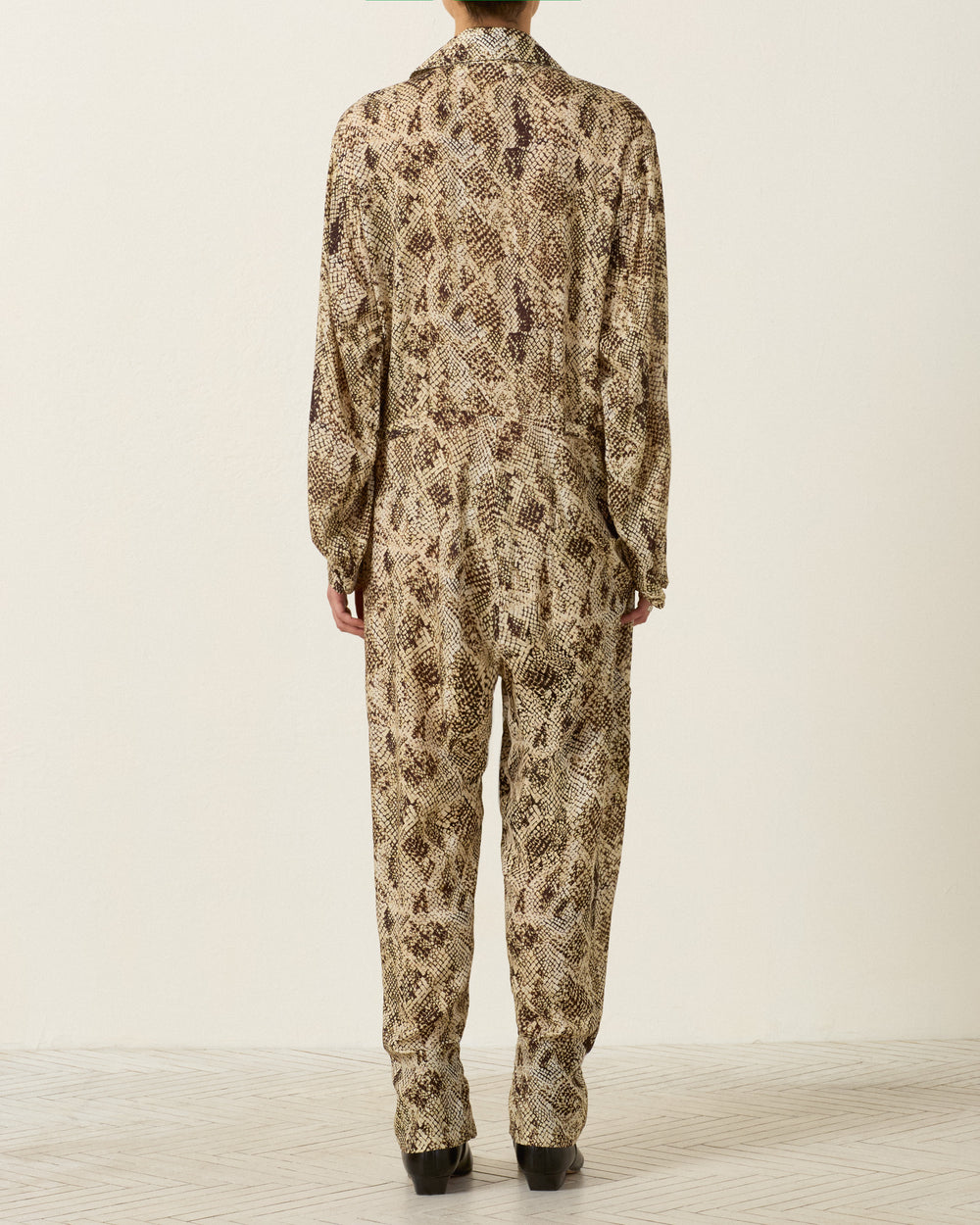 A person stands with their back to the camera, wearing the Miles Lemon Snakeskin Jumpsuit—a workwear-inspired, long-sleeved jumpsuit in a brown and beige snakeskin print—against a plain light-colored wall and textured floor.