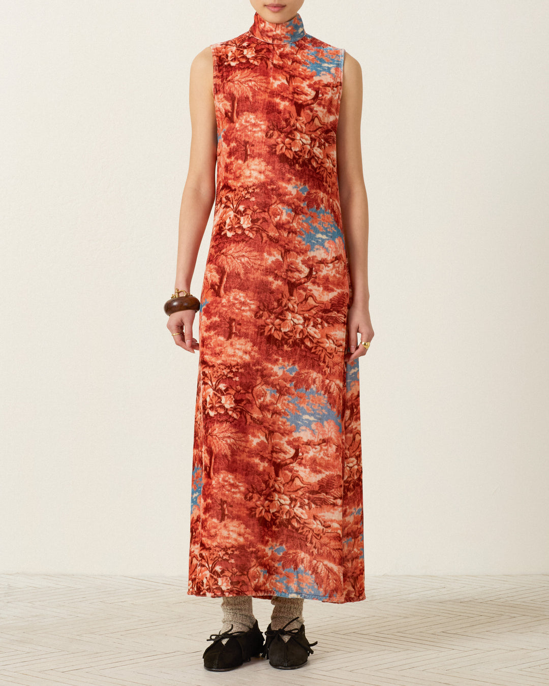 A person wearing the Nadia Heritage Canopy Dress—a sleeveless, high-neck, ankle-length red and blue floral viscose-silk blend dress—paired with brown bracelets, socks, and black shoes, standing against a plain background.