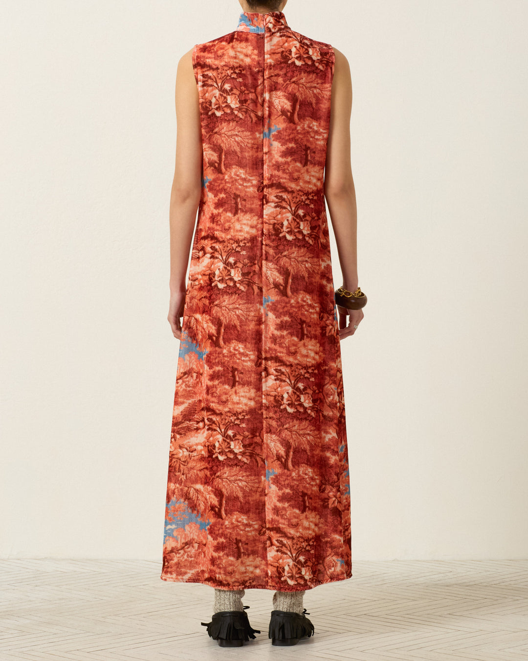 A person stands with their back to the camera, wearing the Nadia Heritage Canopy Dress—a sleeveless, long red patterned piece crafted from a luxurious viscose-silk blend—paired with socks and black fringed shoes. The background is plain, and the floor is light-colored.