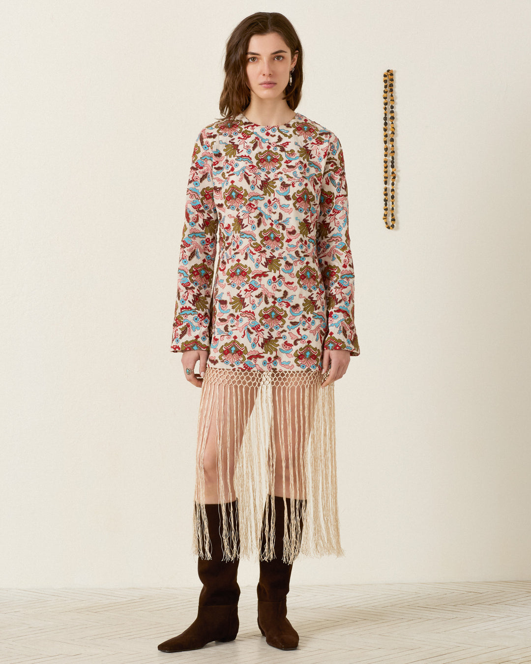 A woman stands against a plain wall wearing the Narene Kilim Emblem Dress—its floral long sleeves, fringe detailing, and delicate hand-beading create intricate texture. She completes the look with black boots, while a beaded decoration hangs on the wall beside her.
