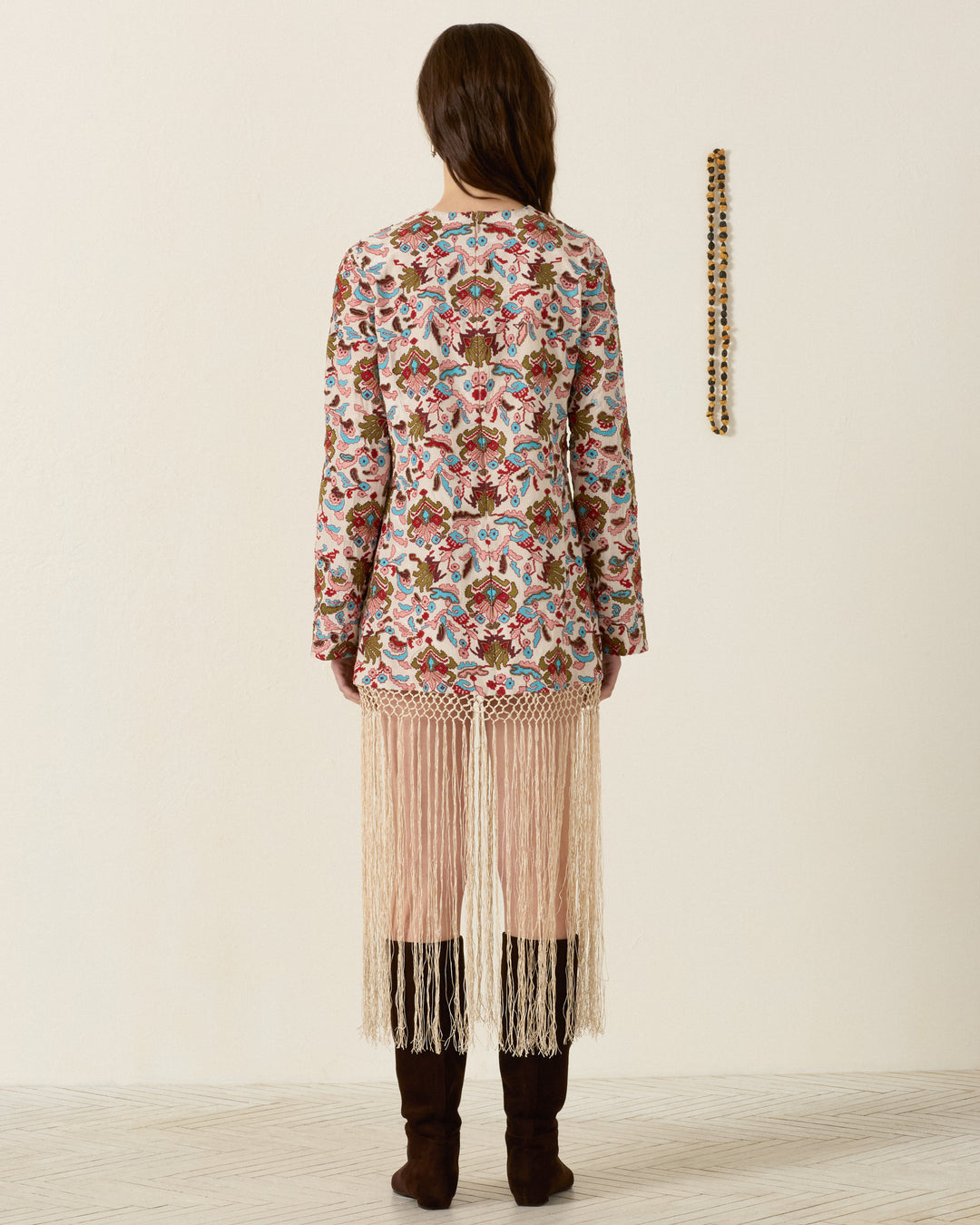A woman stands with her back to the camera, wearing the Narene Kilim Emblem Dress—adorned with floral patterns and long fringe—paired with brown boots, beside a beaded necklace featuring hand beading and kilim-inspired motifs hanging on a cream-colored wall.