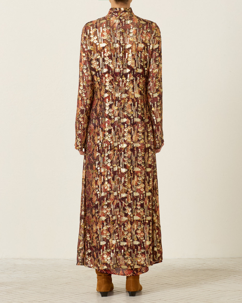 A person stands facing away, wearing the long-sleeved, ankle-length Nia Topaz Snakeskin Dress paired with brown boots.