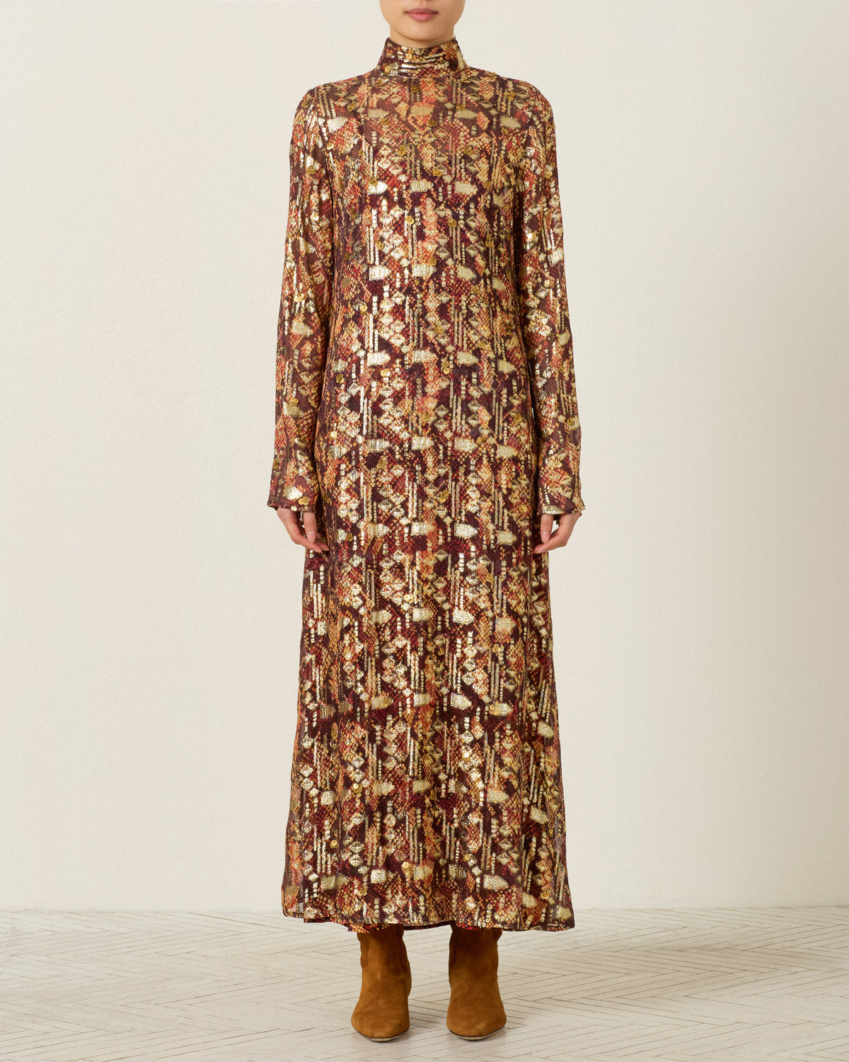 A person wearing the Nia Topaz Snakeskin Dress—a long-sleeved, high-neck, ankle-length hand-embellished piece featuring a gold and brown geometric pattern—paired with brown boots, stands against a plain background.