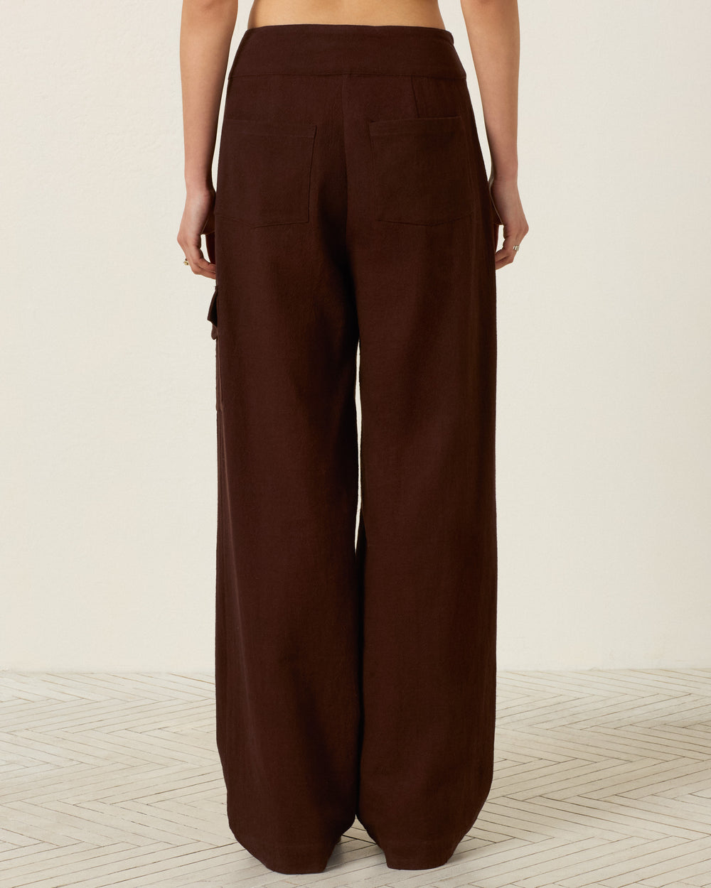A person wearing the Painter Woodland Pant in dark brown, featuring a high-waisted, wide-leg silhouette with two back pockets and distinctive cargo pocket detailing, stands on a light patterned floor against a plain white wall.