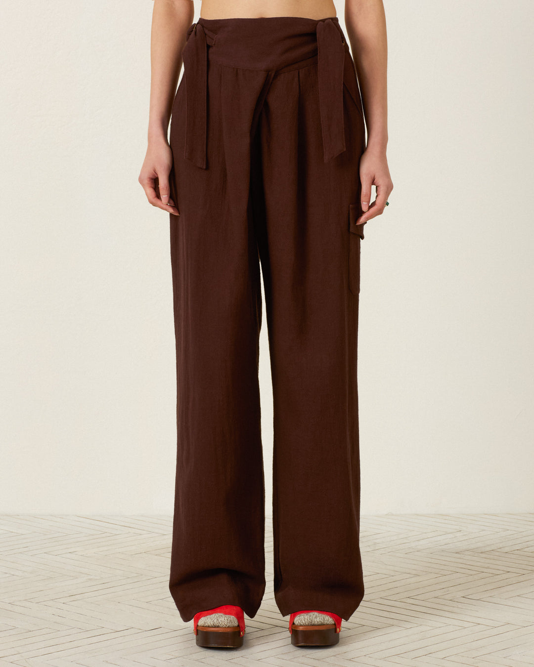 Wearing the Painter Woodland Pant—high-waisted, wide-leg field pants with adjustable side tabs and a sash belt—paired with wooden platform sandals featuring red straps, the person stands against a light background.