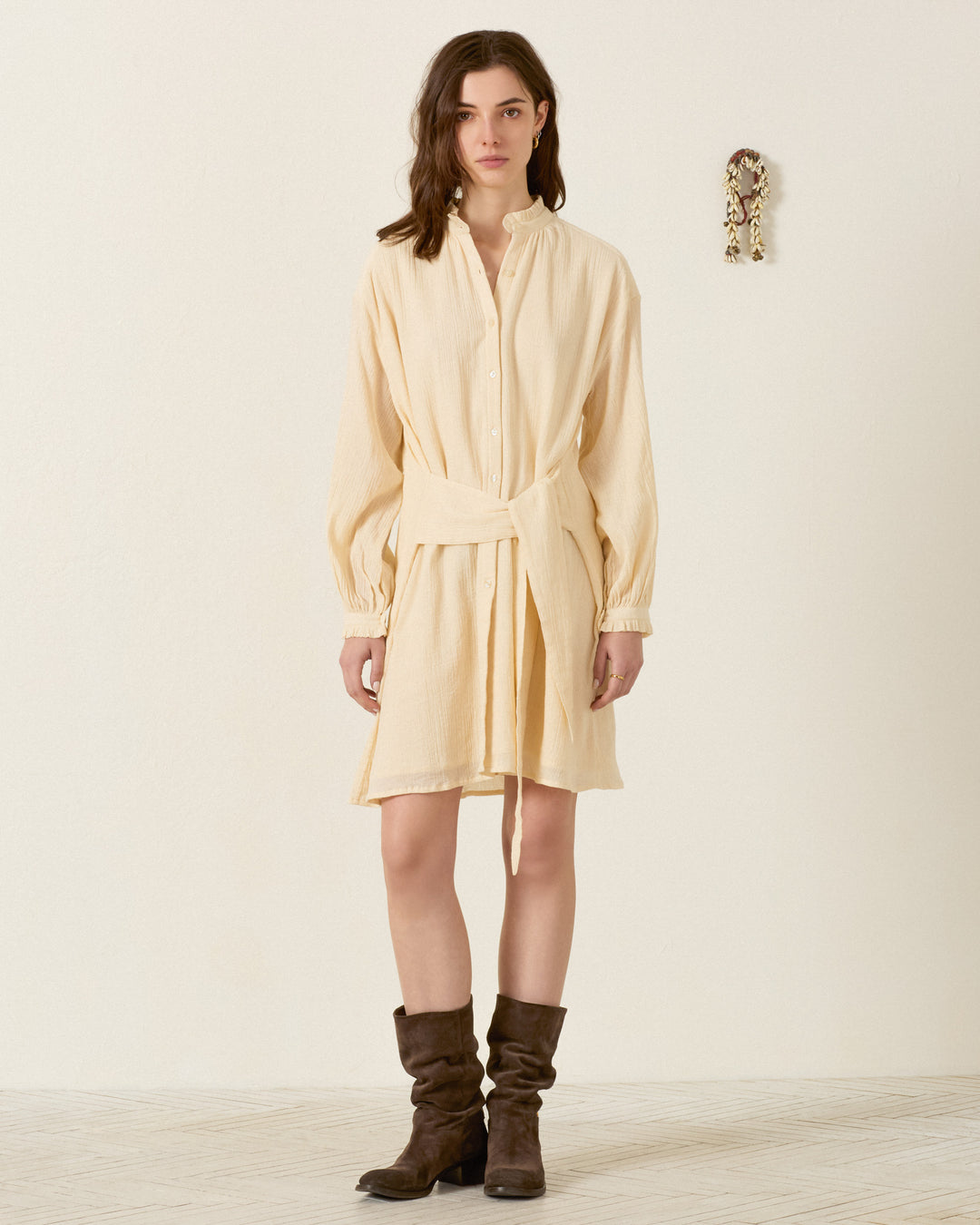 A woman stands indoors against a plain wall, wearing the Parvati Prairie Tint Dress—a cream-colored cotton shirt dress with waist ties—paired with brown knee-high boots.