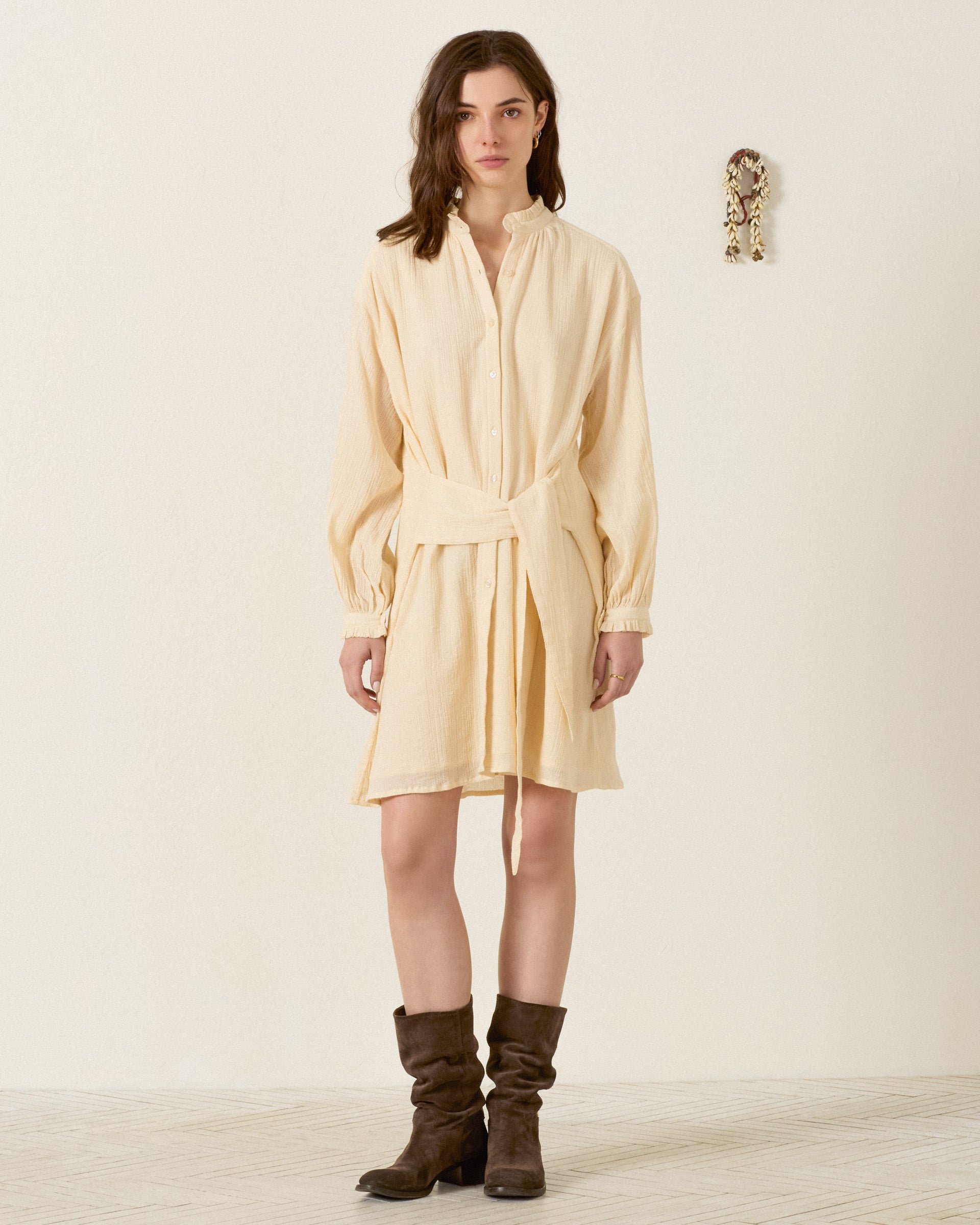 A woman stands indoors against a plain wall, wearing the Parvati Prairie Tint Dress—a cream-colored cotton shirt dress with waist ties—paired with brown knee-high boots.