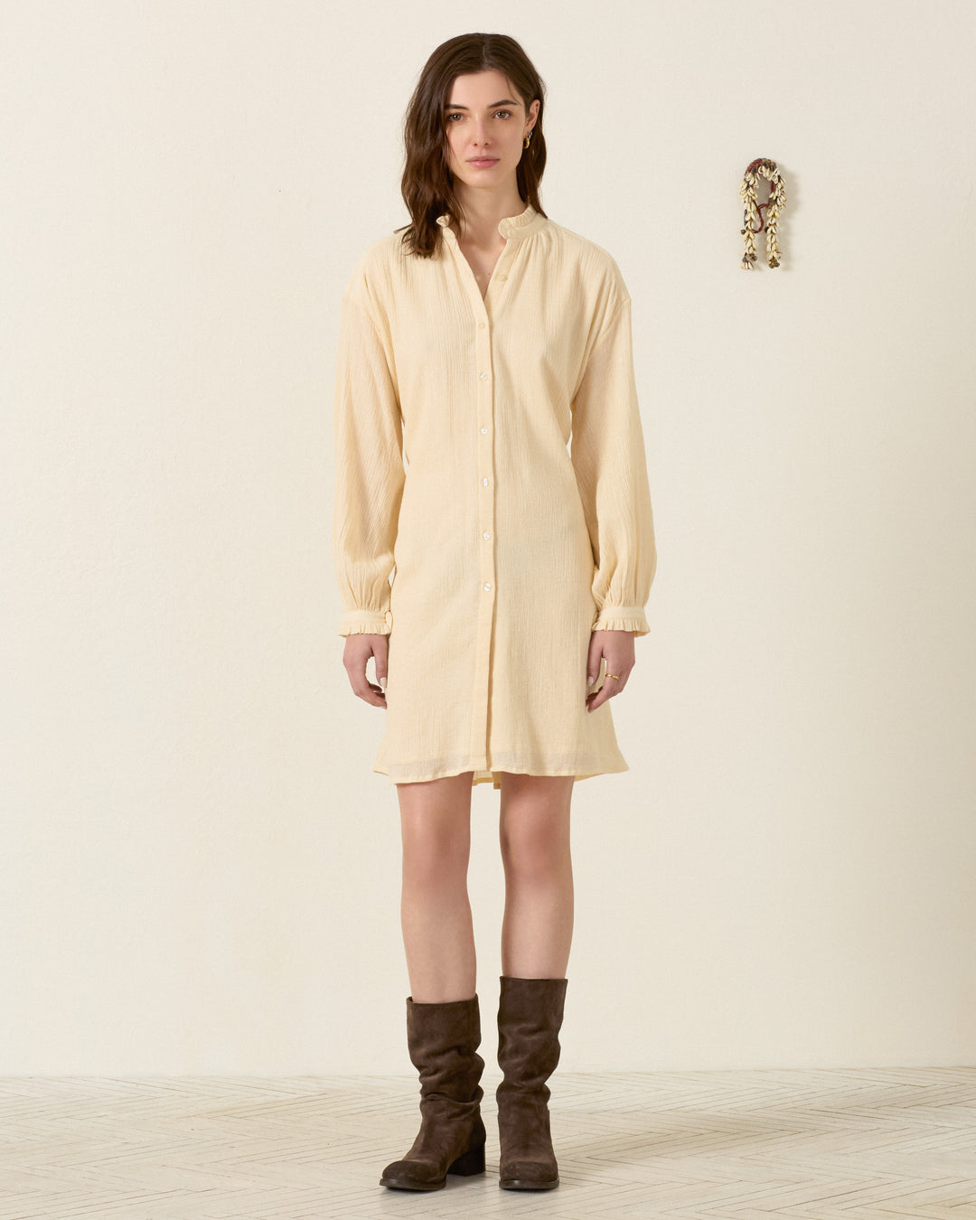 A woman stands indoors facing forward against a plain wall with a small decorative item, wearing the Parvati Prairie Tint Dress—a light beige cotton dobby shirt dress featuring a subtle ruffled collar—paired with dark brown boots.