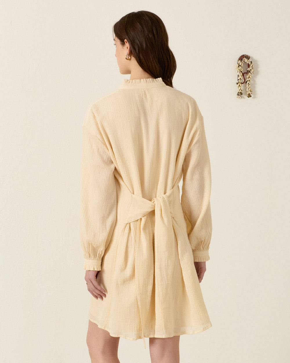 A woman stands with her back to the camera, wearing the Parvati Prairie Tint Dress—a long-sleeved, light beige cotton dobby shirt dress with a waist tie—against a plain light-colored wall adorned with a small decorative item.