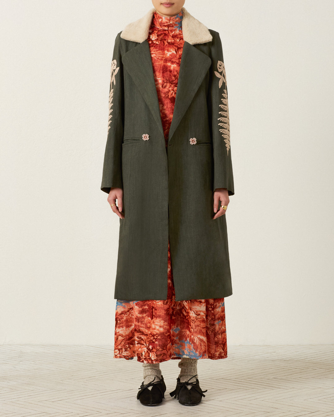 A person wears the Petunia Grassland Fern Coat, a long dark green wool-linen topcoat with hand-embroidered details and an Italian wool fleece collar, layered over an orange patterned dress, paired with patterned socks and black shoes, standing against a plain background.