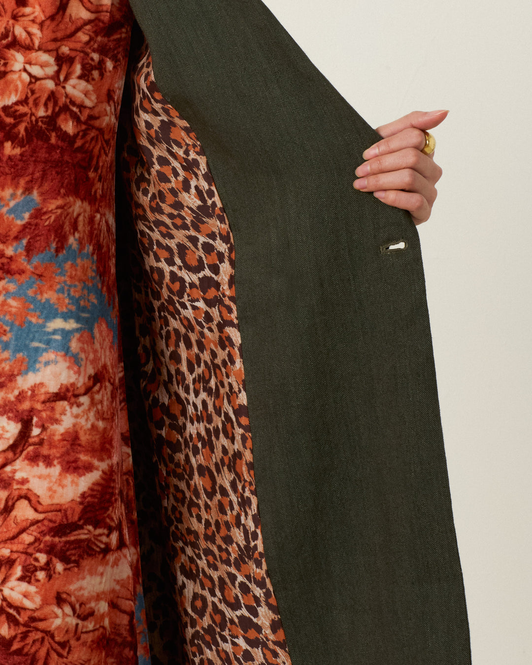 A person holds open the Petunia Grassland Fern Coat—a dark green wool-linen topcoat—revealing its leopard print lining. The coat is finished with an Italian wool fleece collar, and underneath, the person wears a red, orange, and blue floral patterned garment.