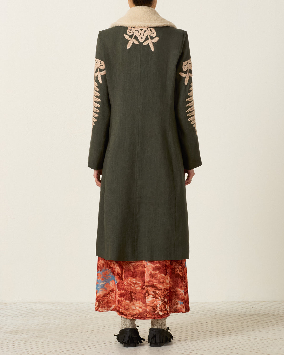 A person stands facing away, wearing the Petunia Grassland Fern Coat—a hand-embroidered long dark coat with an Italian wool fleece collar—over a red and blue patterned skirt, paired with black shoes featuring fringe detail.
