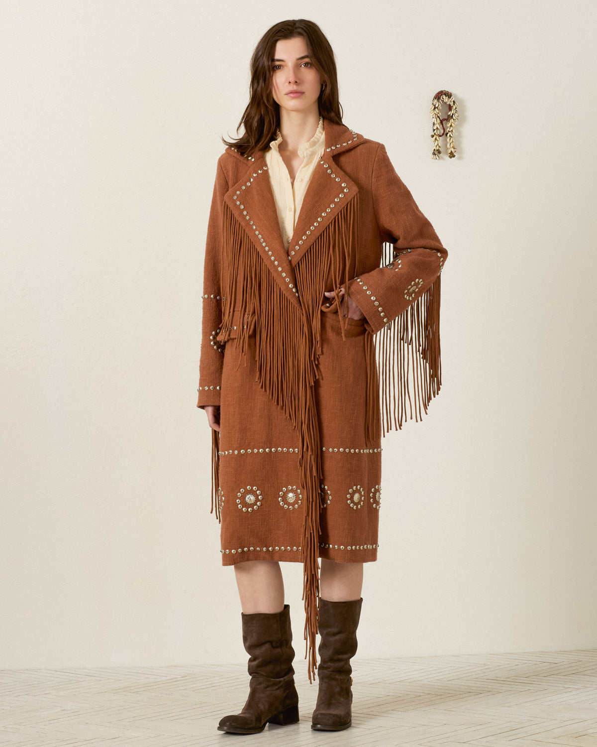 A woman stands indoors wearing the Petunia Fawn Stud Coat—a Western-inspired brown fringed layer with studded designs—paired with a cream shirt and brown boots.