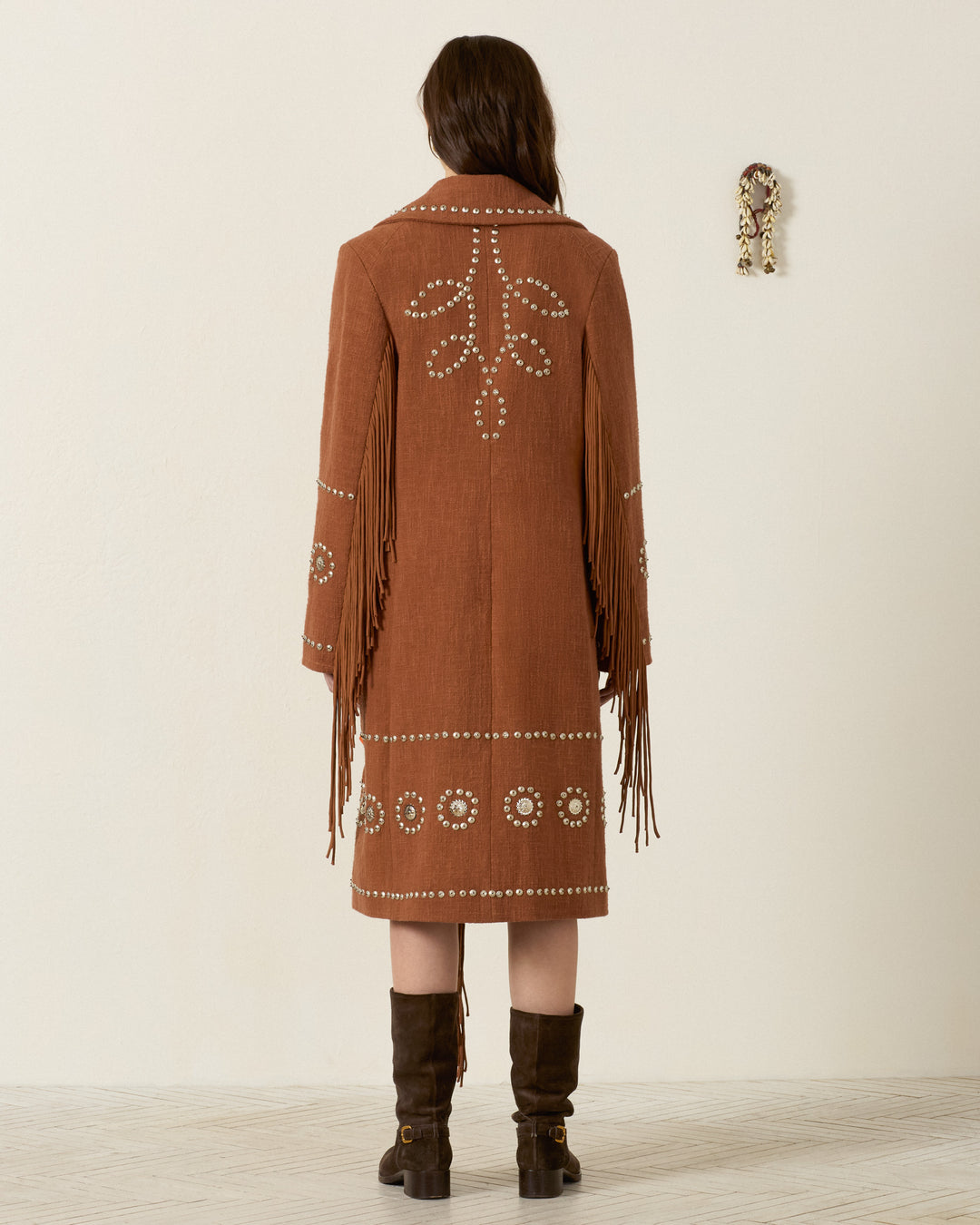 Wearing the Petunia Fawn Stud Coat—a brown, knee-length khadi cotton coat featuring decorative white patterns and fringe sleeves—a person stands facing away, paired with dark brown boots.