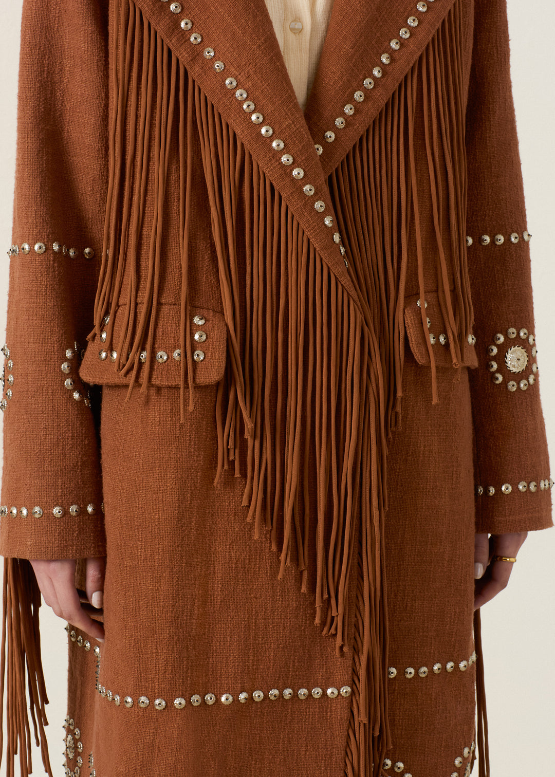 Wearing the Petunia Fawn Stud Coat, a Western-inspired brown outerwear piece adorned with long fringe details and decorative silver embellishments on the sleeves, front, and pockets. This statement coat features a hand-embellished finish and is styled over a beige shirt for a look that merges classic style with modern flair.