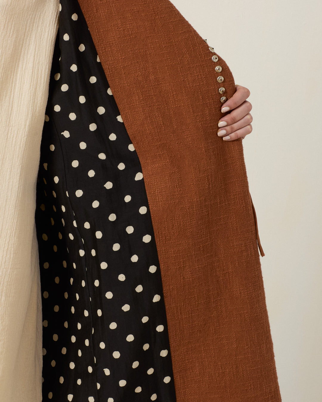 A person holds open the Petunia Fawn Stud Coat, a rust-colored textured Khadi cotton coat featuring a black and white polka dot lining and a row of small buttons along the inside edge.