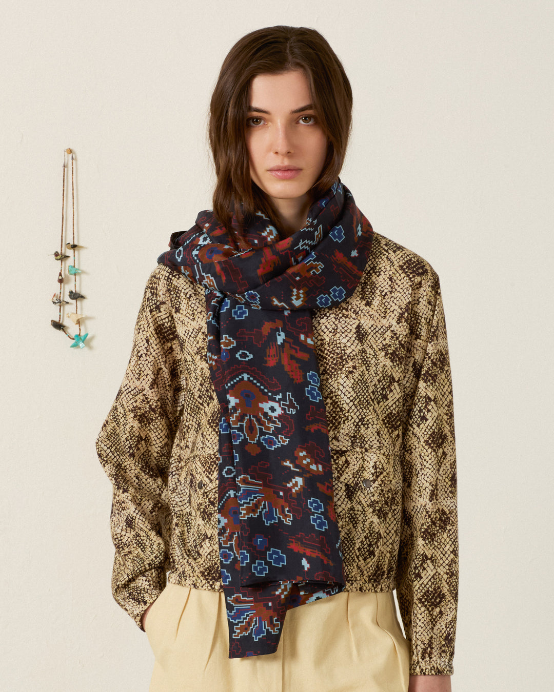 A woman stands against a plain wall wearing a beige snakeskin-patterned shirt, cream pants, and the Pietra Indigo Kilim Silk Scarf with dark patterns. A small hanging decor with beads is visible on the wall beside her.