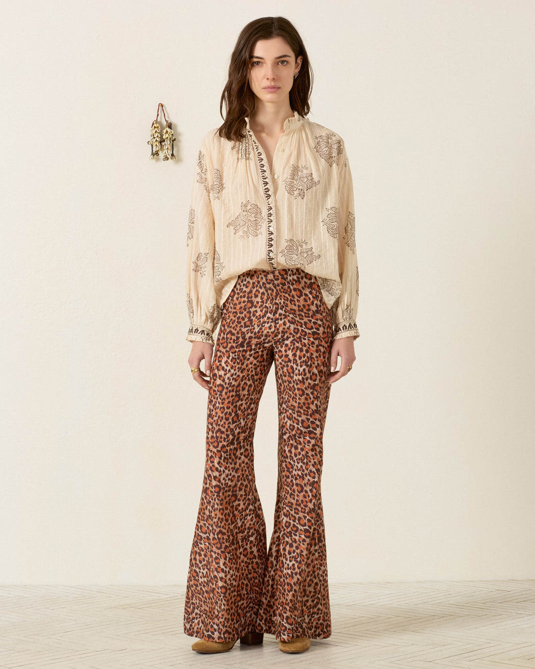 A woman stands indoors against a plain light wall with a small hanging decor item, wearing the Farah Leopard Trek Pant—a high-waisted, bell-bottom flare pant in a bold leopard print—paired with a cream embroidered blouse and tan shoes.