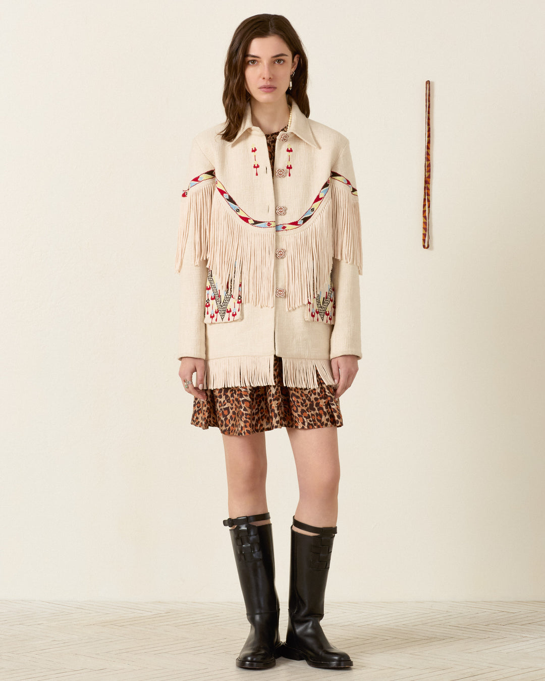 A woman stands indoors wearing the Ranger Prairie Fringe Jacket with handcrafted floral buttons, layered over a leopard print skirt and tall black boots. A wooden rod is mounted vertically on the plain wall behind her.