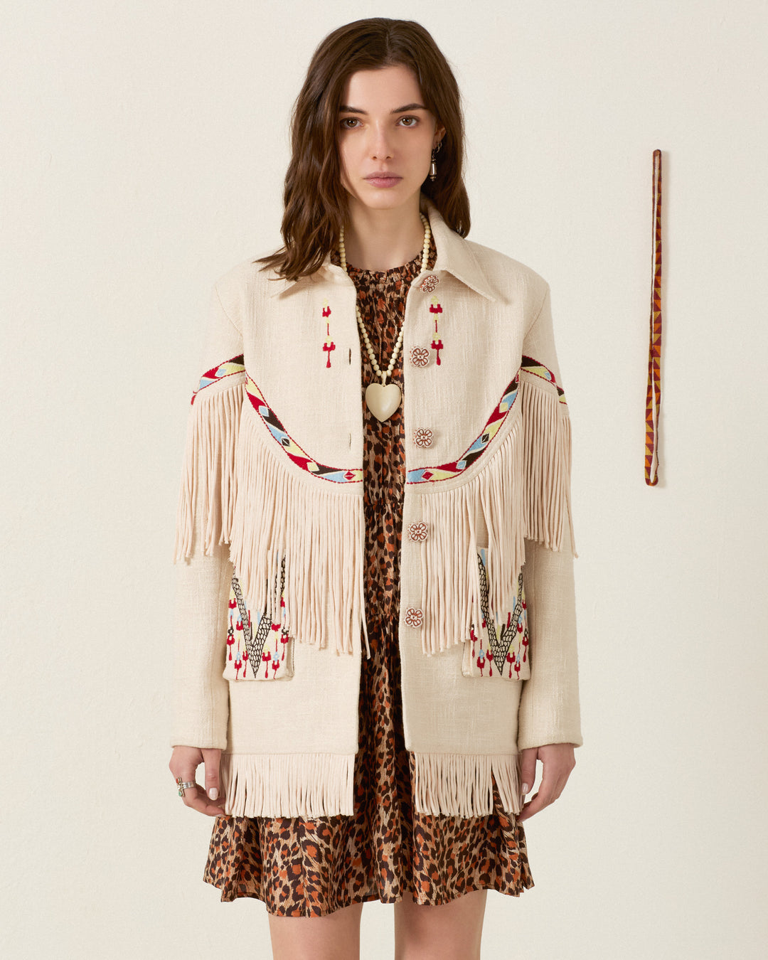 A woman stands against a plain wall, wearing the Ranger Prairie Fringe Jacket—an embroidered piece with handcrafted floral buttons—layered over a leopard print dress; she also accessorizes with a beaded necklace and holds a long stick.