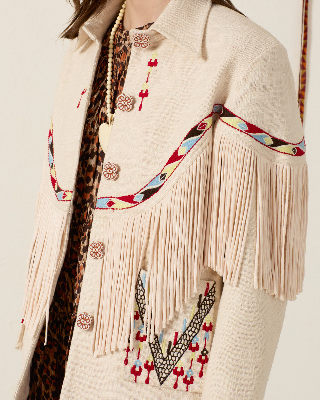 The Ranger Prairie Fringe Jacket is a cream-colored Khadi cotton piece featuring embroidered patterns, handcrafted floral buttons, fringe detailing on the chest and sleeves, and button closures; a patterned shirt is partially visible underneath.