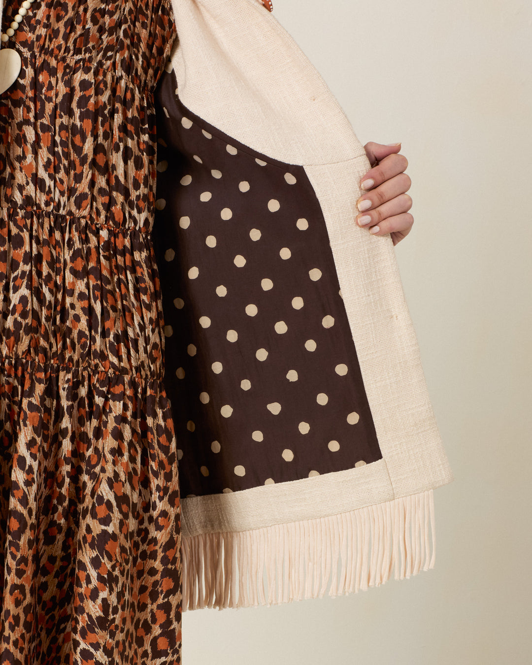 A person holds open the Ranger Prairie Fringe Jacket, showcasing its cream-colored Khadi cotton with delicate fringe detailing and a brown polka dot lining. The jacket is styled over a brown and orange leopard print dress, and features handcrafted floral buttons that add a unique touch to the ensemble.