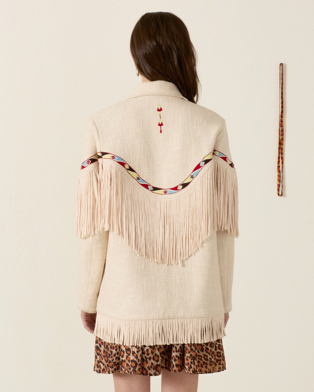 A woman with long brown hair, seen from behind, wears the Ranger Prairie Fringe Jacket—an embroidered piece with fringe and patterned trim—over a leopard print skirt. A decorated stick hangs on the wall to the right.