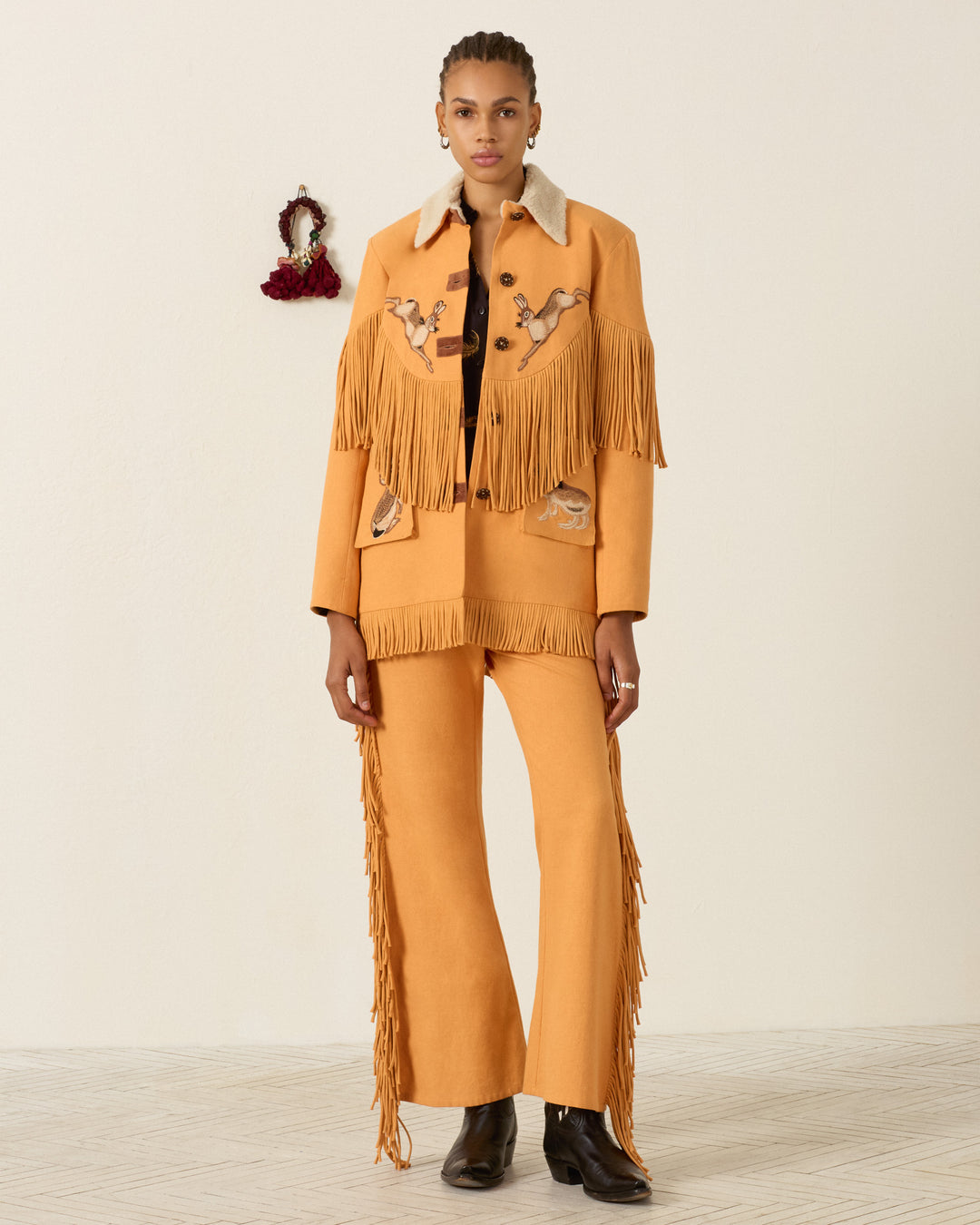 A person stands against a plain wall, wearing a tan fringed jacket with Western-inspired details, matching it with the Bunny Sunset Fringe Pant and black boots.