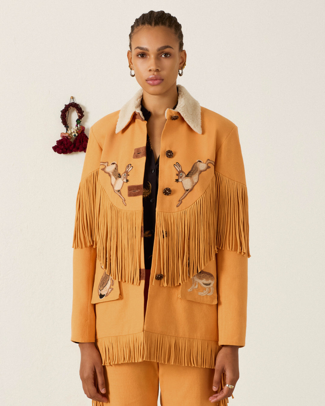 A woman stands against a plain light background wearing the Ranger Sunset Hare Jacket, a mustard yellow fringed jacket crafted from sumptuous Italian wool fleece and adorned with hand-embroidered animals.