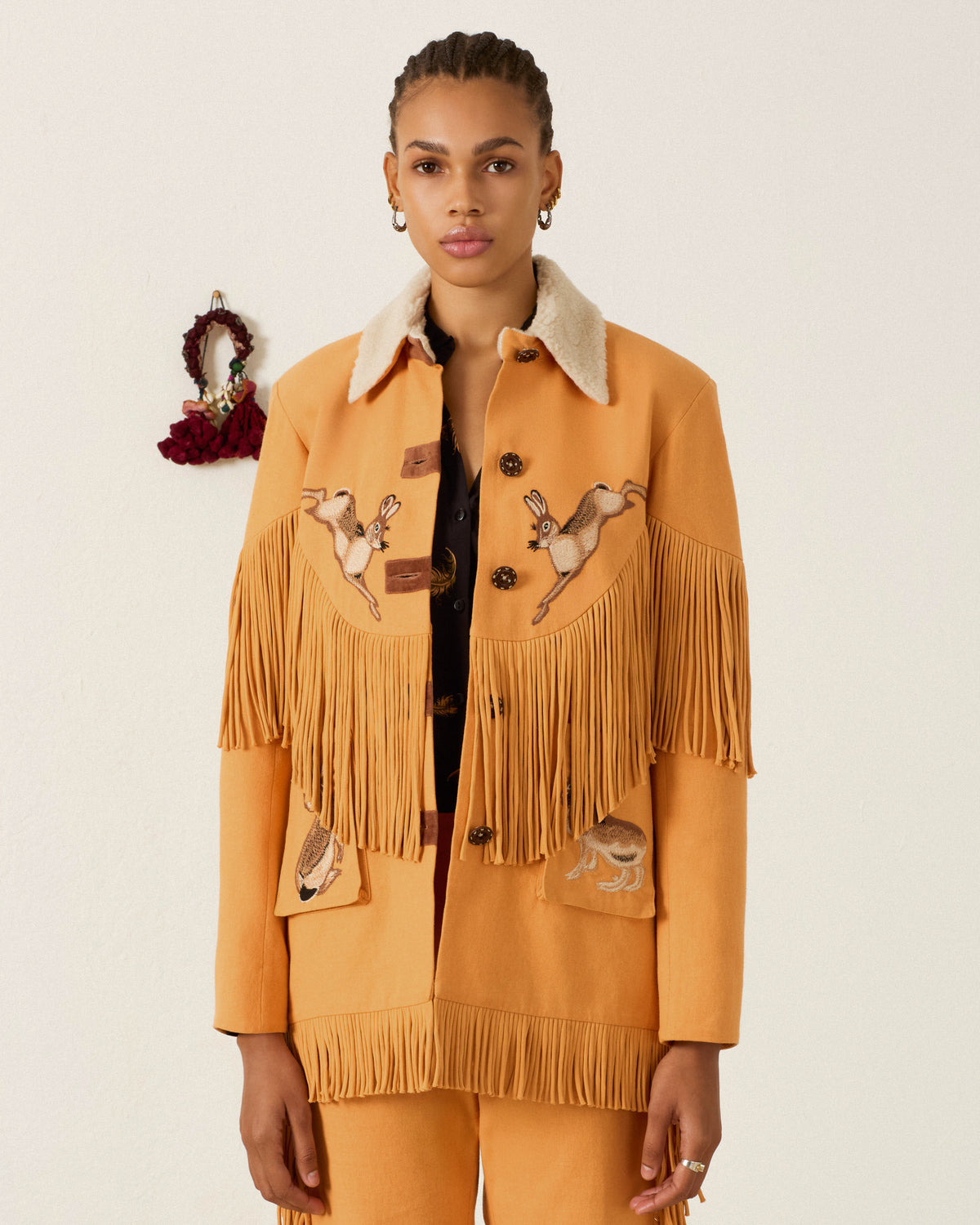 A woman stands against a plain light background wearing the Ranger Sunset Hare Jacket, a mustard yellow fringed jacket crafted from sumptuous Italian wool fleece and adorned with hand-embroidered animals.