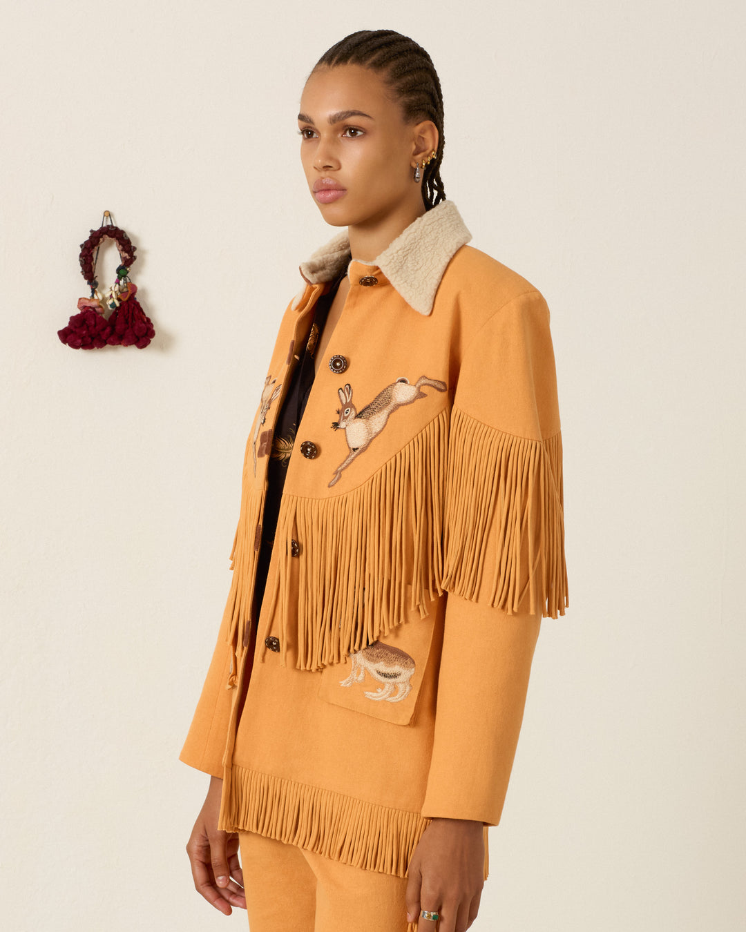 A person wearing the Ranger Sunset Hare Jacket—an orange, hand-embroidered piece featuring animal motifs, a shearling collar, and crafted from Italian wool fleece—stands indoors near a small wall decoration.
