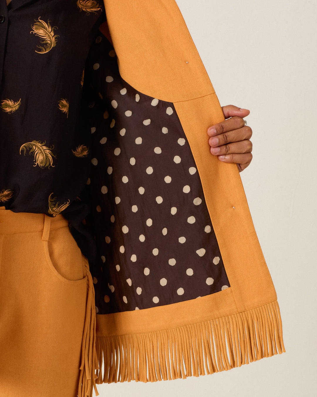 A person holds open the Ranger Sunset Hare Jacket—a mustard yellow, fringed piece made from luxurious Italian wool fleece—revealing its black lining with white polka dots. The ensemble is completed with a black shirt featuring yellow paisley patterns and coordinating shorts.