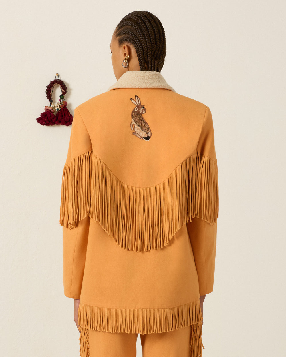 A person faces away from the camera wearing the Ranger Sunset Hare Jacket, a tan fringed piece featuring a hand-embroidered jackrabbit design on the back. A woven art piece hangs on the wall to the left.