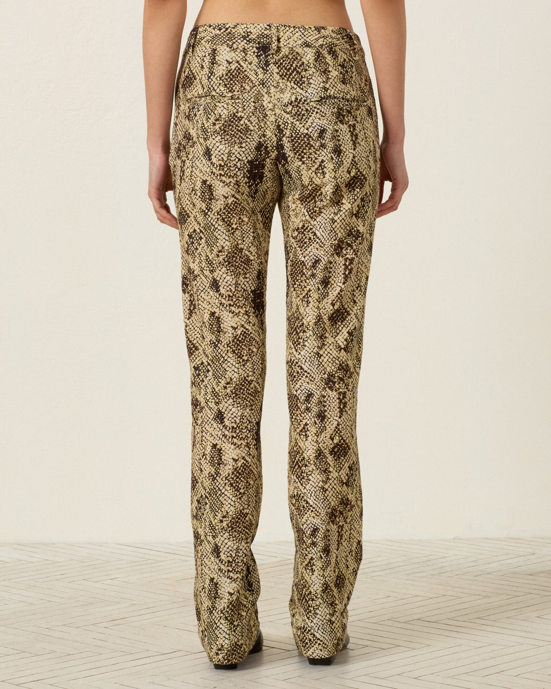 A person stands with their back to the camera, wearing the Razza Lemon Snakeskin Pant—high-waisted, slim-fit pants featuring a beige and brown snakeskin print.