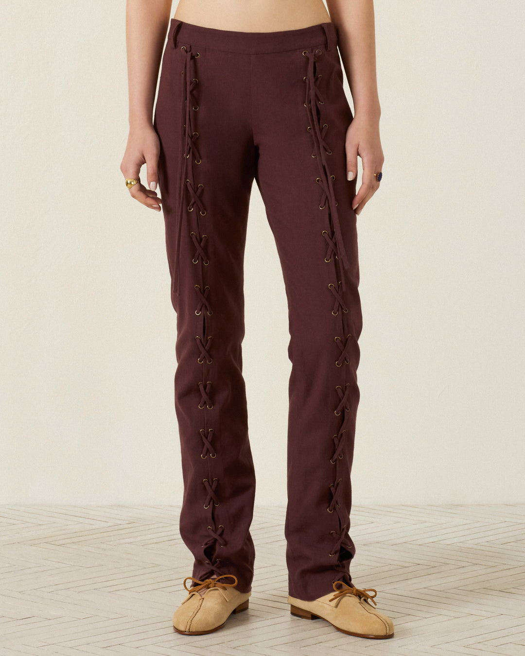 Wearing the Razza Mahogany Pant—crafted from maroon cotton with straight legs, grommeted lace-up ties, side eyelets, and delicate French seams—paired with tan soft shoes, standing on a light patterned floor against a plain background.