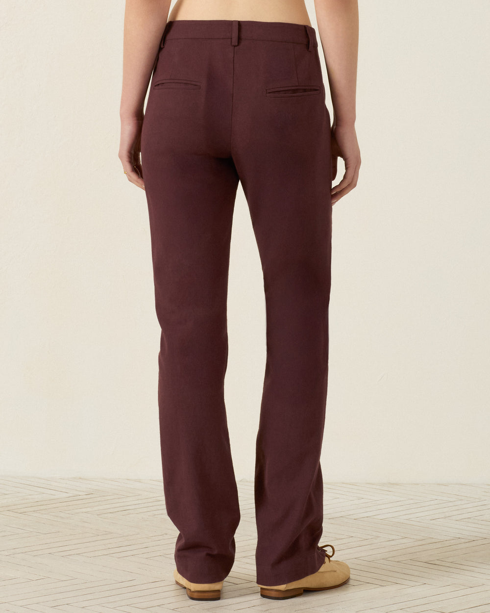 A person is standing facing away, wearing the Razza Mahogany Pant—burgundy cotton straight-leg pants with grommeted lace-up ties—and beige shoes, photographed indoors against a plain light wall and patterned floor.