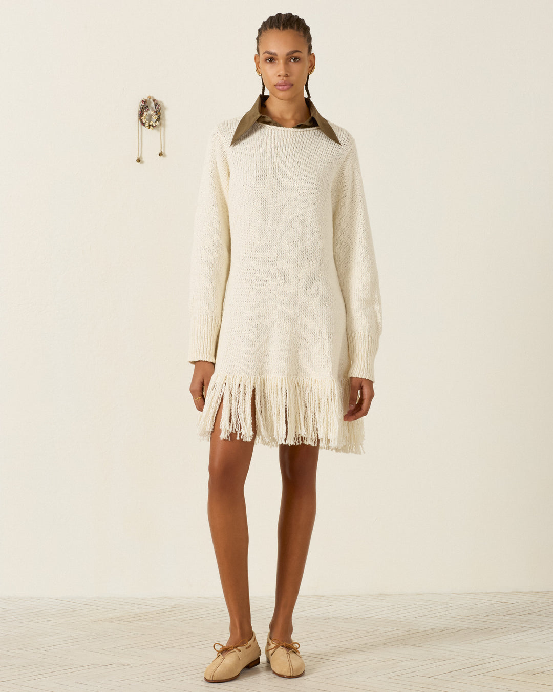 Wearing the Renny Bone Fringe Dress—a long-sleeve, cream-hued hand-knitted sweater dress made from soft Peruvian cotton and finished with fringe at the hem—paired with tan shoes and a large brown collar, a person stands against a plain light wall.