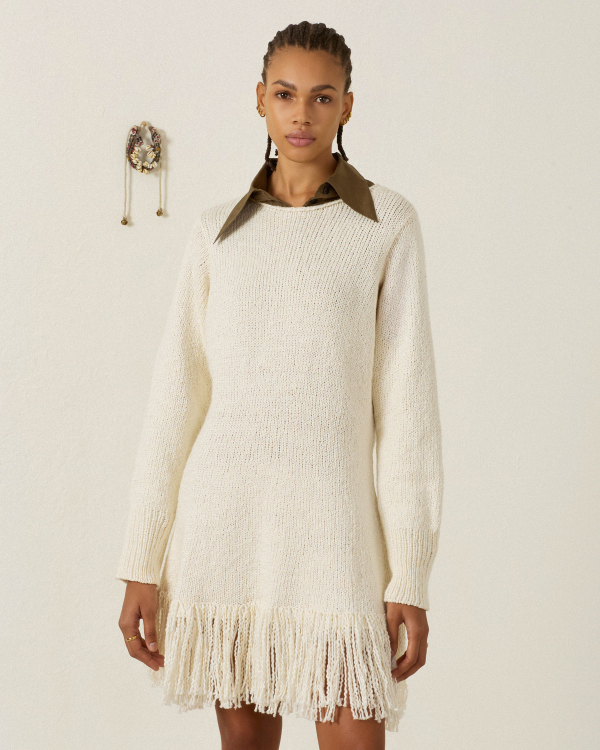 A woman wearing the Renny Bone Fringe Dress—a cream-colored, long-sleeve hand-knitted sweater dress with a fringe hem, crafted from soft Peruvian cotton—stands against a plain off-white wall with a decorative object to her left.