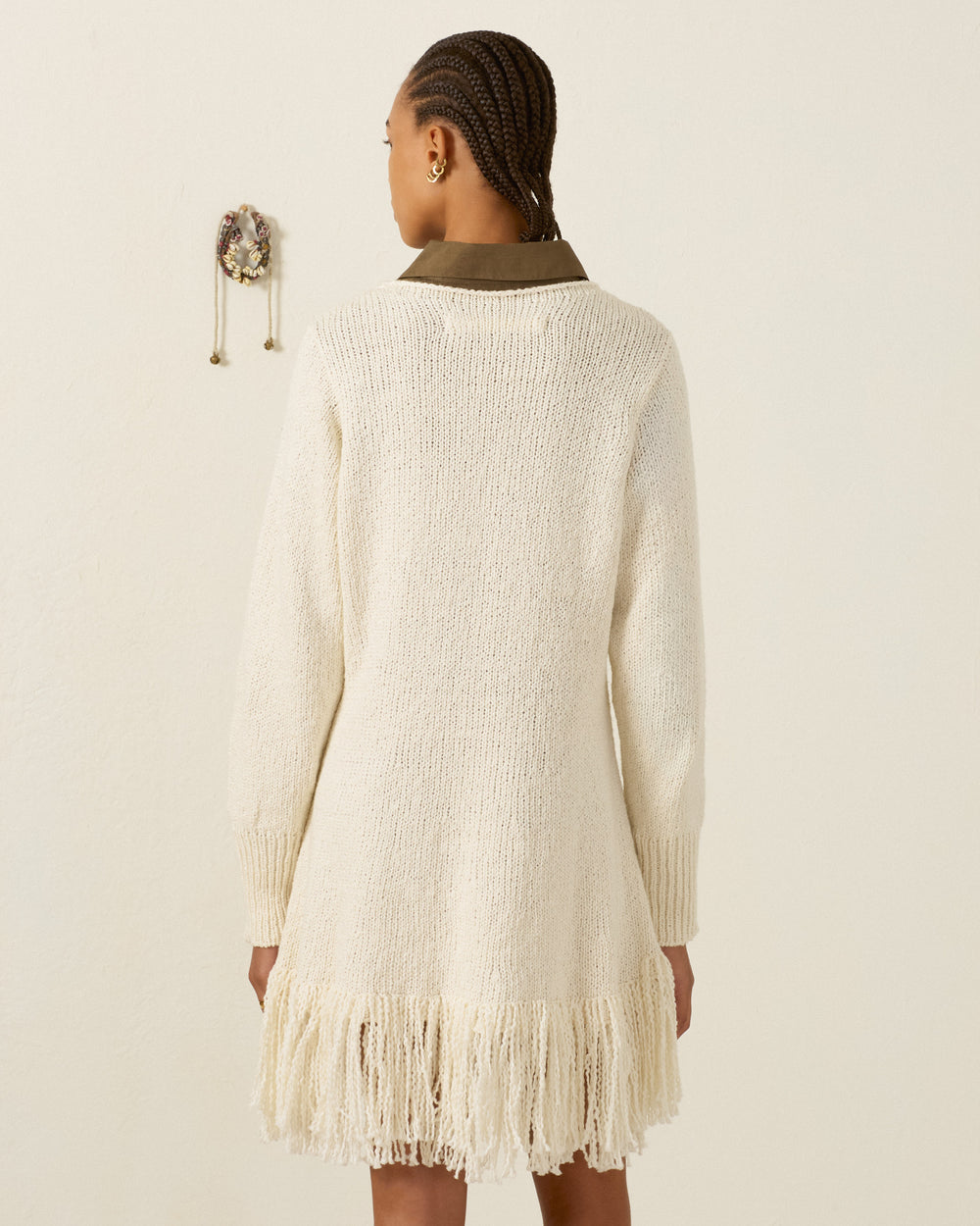 Woman standing with her back to the camera, wearing the artisan-made Renny Bone Fringe Dress— a cream-colored knit piece featuring long sleeves and fringe at the hem.