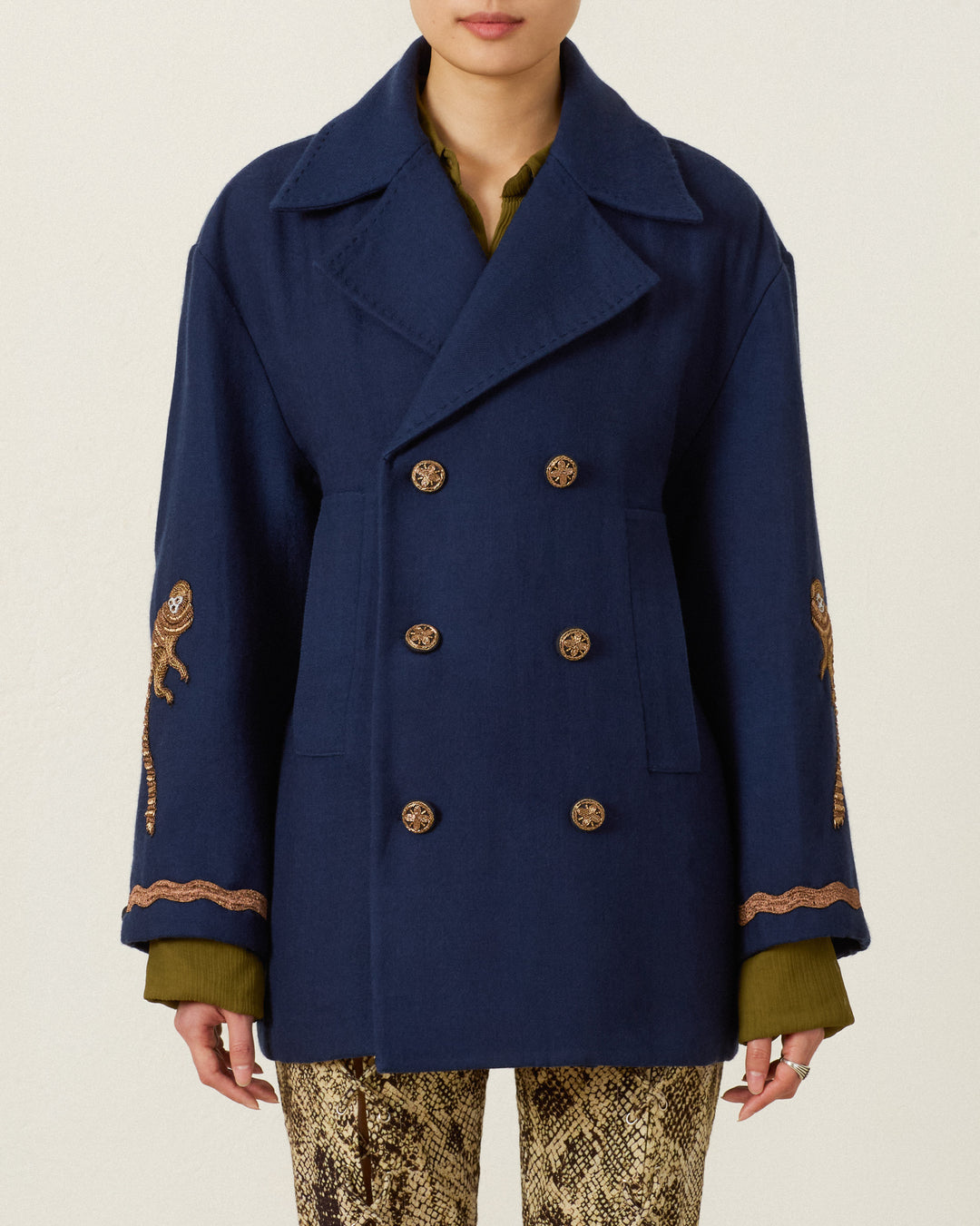 Wearing the Rivoli Midnight Mischief Coat—a navy double-breasted wool peacoat with hand-beaded embroidered designs on the sleeves—over a green shirt and snake print pants, this person stands against a plain light background.
