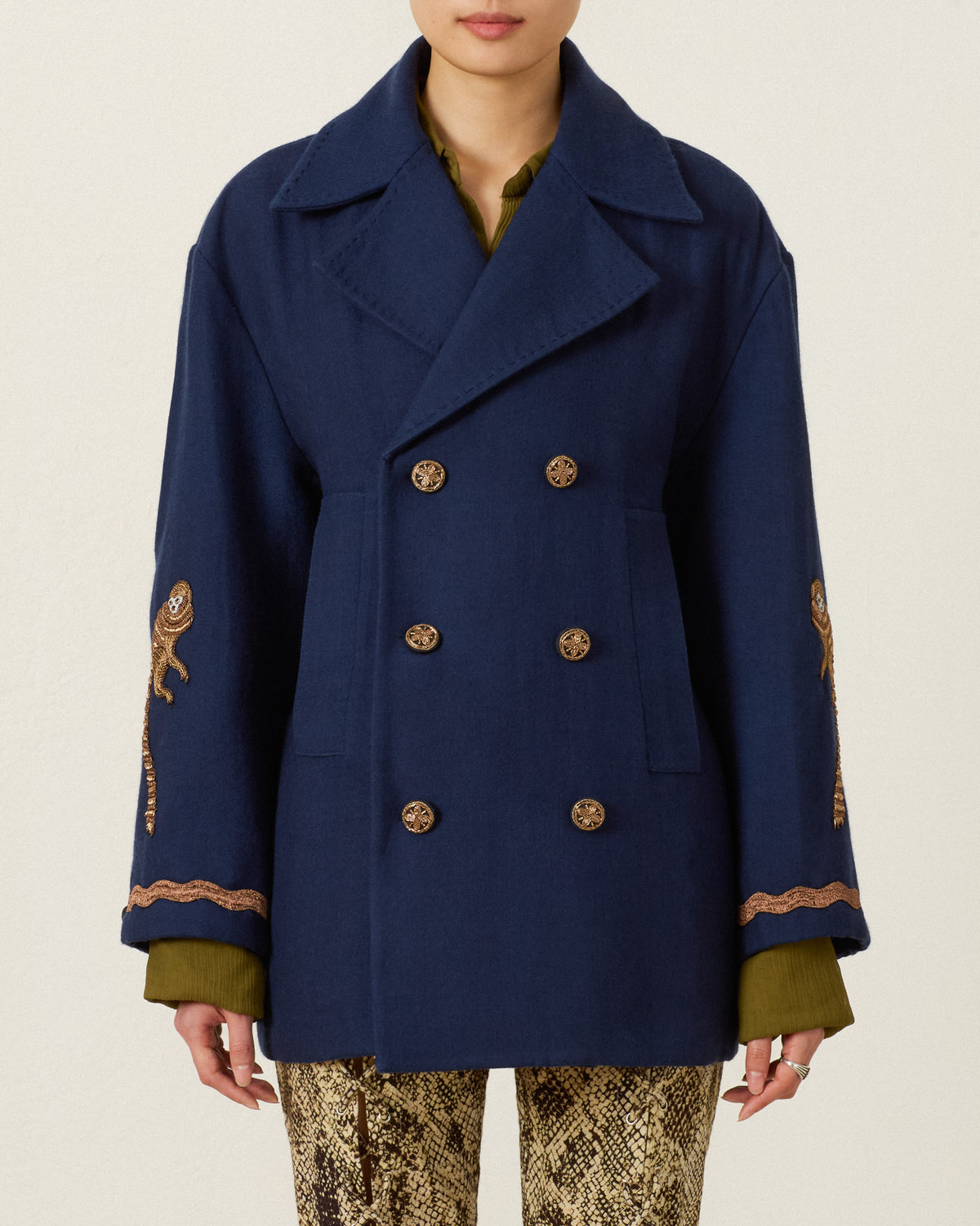 Wearing the Rivoli Midnight Mischief Coat—a navy double-breasted wool peacoat with hand-beaded embroidered designs on the sleeves—over a green shirt and snake print pants, this person stands against a plain light background.