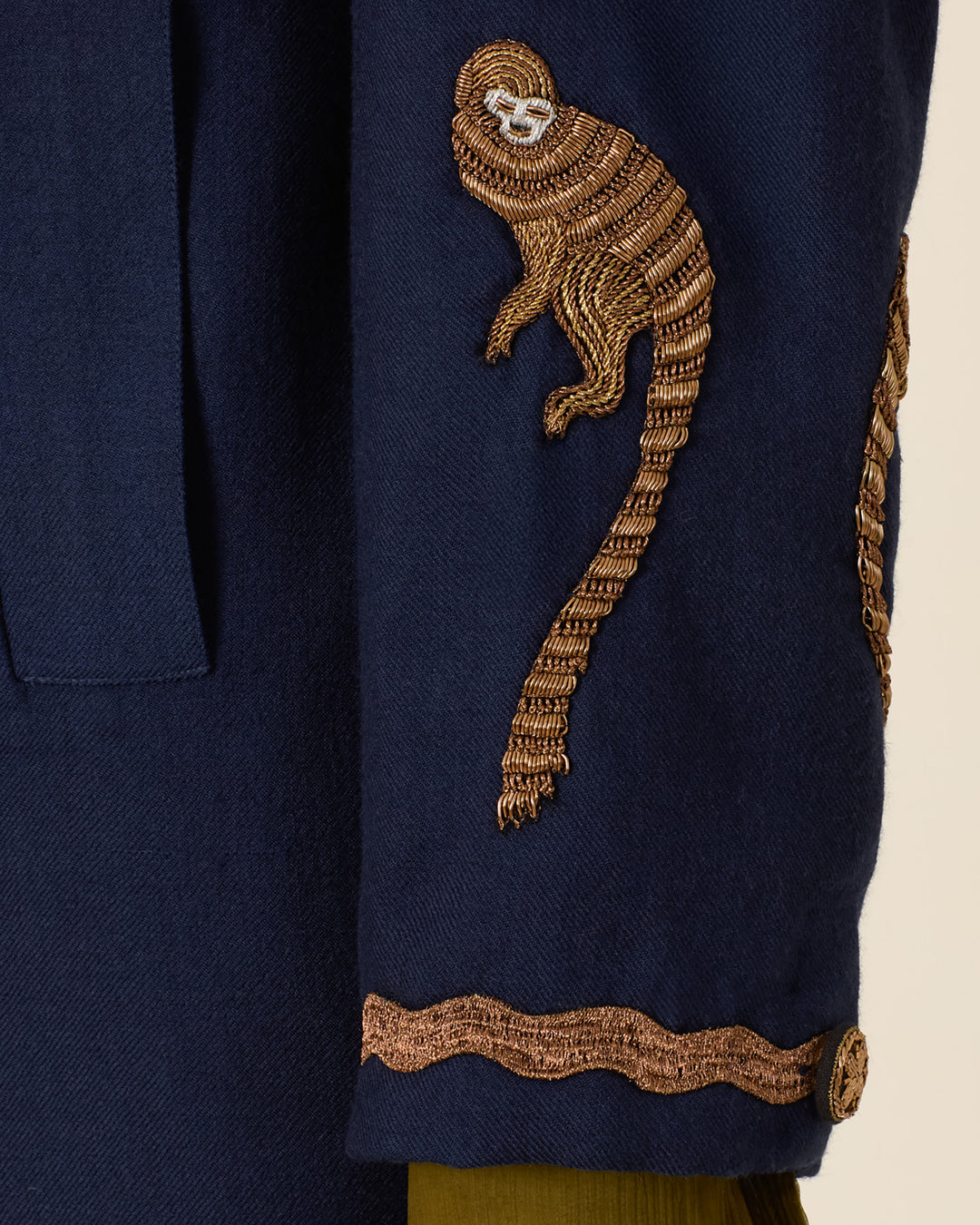 A close-up of a navy blue sleeve on the Rivoli Midnight Mischief Coat, showcasing an embroidered gold monkey and a wavy gold trim near the cuff.